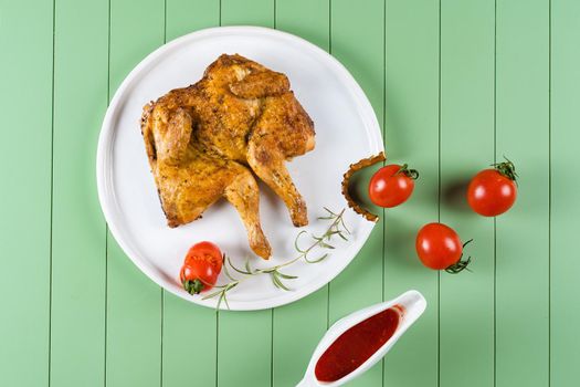 Chicken tobacco with tomato sauce, rosemary and tomatoes on a beautiful white plate on a green background. Grilled chicken.