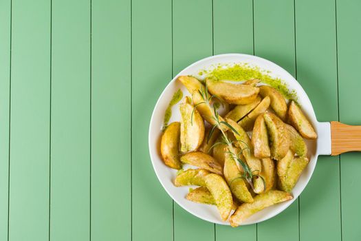 Baked potatoes with green sauce and rosemary on a white plate in the form of a frying pan. Fried potatoes with pesto sauce on a green table.