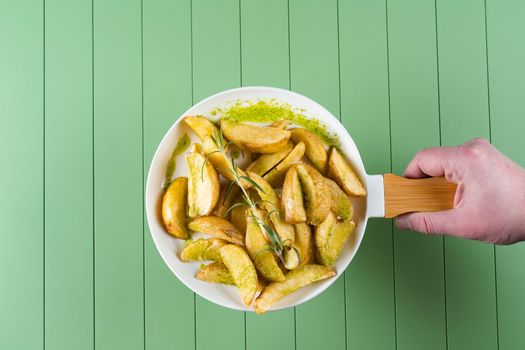 Baked potatoes with green sauce and rosemary on a white plate in the form of a frying pan. Fried potatoes with pesto sauce on a green table.