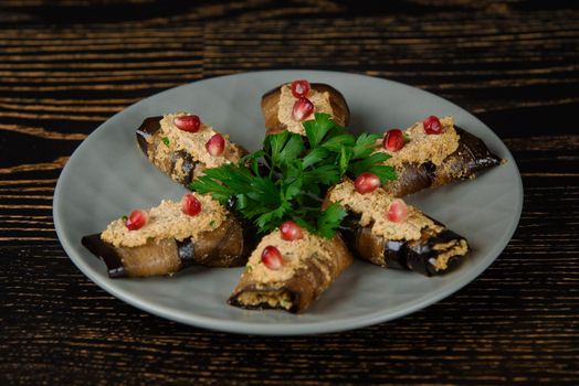 Eggplant rolls decorated with parsley and pomegranate. Georgian appetizer on a gray plate on a dark wooden table.