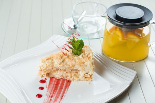 Napoleon cake decorated with a spiral of red chocolate, mint and berry jam on a white plate against a background of orange tea in a teapot and a transparent cup. Layered cake with pastry cream.