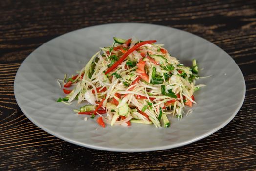 Salad with cabbage, cucumber, tomato, pepper, greens on a gray plate on a dark wooden table. Vegetable salad with oil.