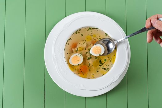 Chicken broth with meat, carrots, herbs and quail egg in a white plate on a green background