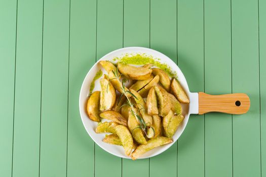 Baked potatoes with green sauce and rosemary on a white plate in the form of a frying pan. Fried potatoes with pesto sauce on a green table.