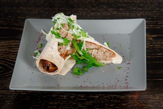 Lula kebab roll in pita bread garnished with onions and cilantro leaves on a gray square plate on a dark wooden table. Georgian dish.