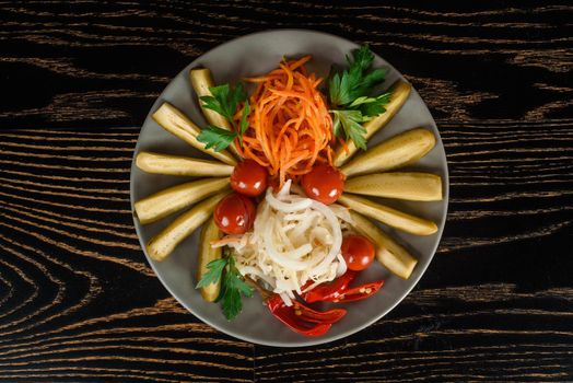 Assortment of pickles, cherry tomatoes, chili with carrots, onions and parsley on a gray plate on a dark wooden table. Top view.