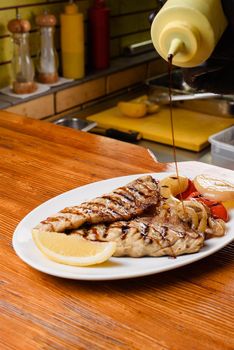 Chicken fillet and grilled vegetables with lemon on a white plate on a wooden table. Pouring sauce onto a dish.