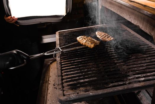 Grilling chicken fillets with metal tongs. The cook prepares a barbecue dish.