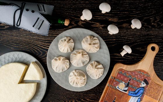 Khinkali on a gray plate, cheese on the background of a bottle in the Georgian style, mushrooms, a board with a thematic pattern on a dark wooden table. Top view