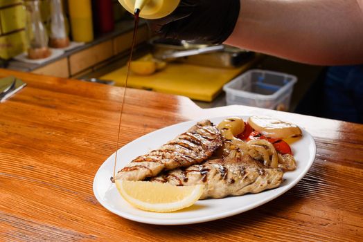 Chicken fillet and grilled vegetables with lemon on a white plate on a wooden table. Pouring sauce onto a dish.