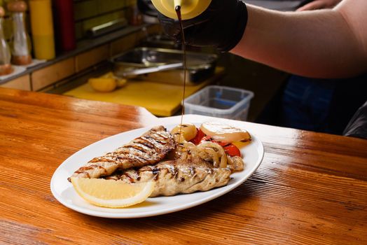 Chicken fillet and grilled vegetables with lemon on a white plate on a wooden table. Pouring sauce onto a dish.