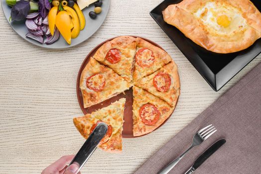 Khachapuri with cheese and tomatoes, egg, slices of vegetables, basil on a white wooden table. Serving. View from above