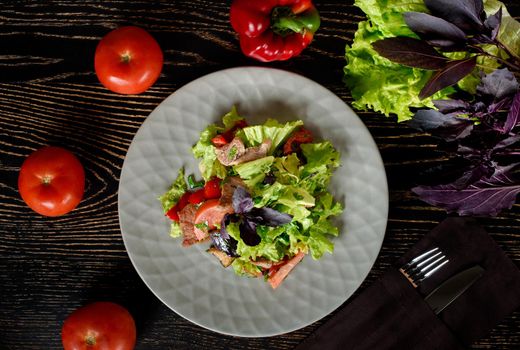 Salad with meat, lettuce, tomatoes, peppers, basil, olive oil on a gray plate on a dark wooden table. Garnished with whole tomatoes, peppers, lettuce, blue basil.