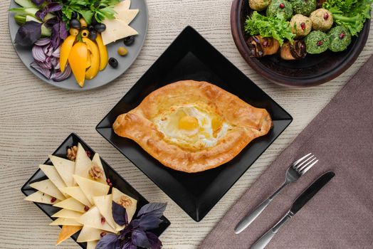 Khachapuri with cheese and tomatoes, egg, slices of vegetables, basil on a white wooden table. Serving. View from above