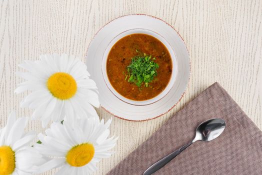 Tomato soup with herbs on a white plate on a light wooden table against a background of chamomile flowers.