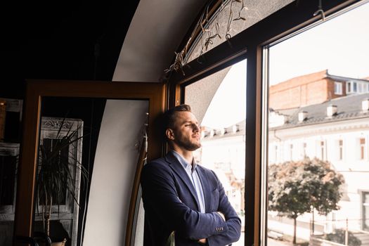 Handsome male model is posing in cafe. Bearded man weared suit stand and smile near window