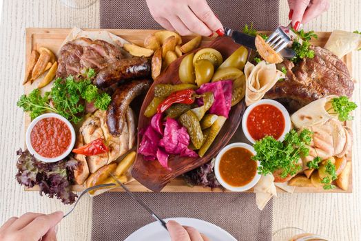 Assortment of fried meat, potatoes, sausages, pickles, tomatoes, peppers, herbs, lavash on a wooden tray on the table. Top view