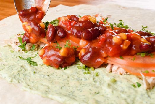 Burrito making process. The cook adds beans and corn in tomato sauce to meat, herbs and tomatoes on pita with sauce. Mexican dish.