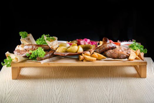 Assortment of fried meat, potatoes, sausages, pickles, tomatoes, peppers, herbs, lavash on a wooden tray on the table.