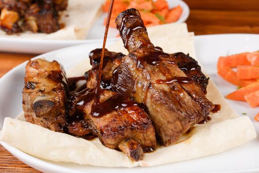 Sliced grilled pork ribs poured with sauce on pita bread with grilled carrots on a white plate on a wooden background.