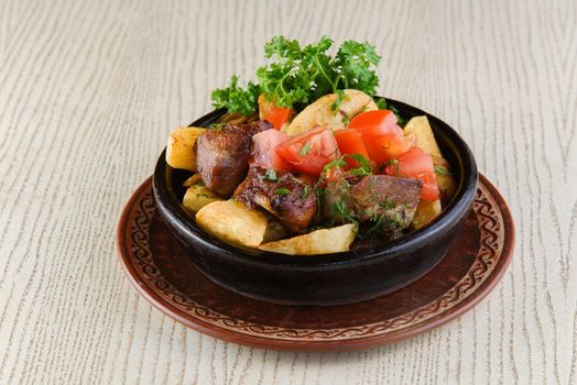 Baked meat with potatoes, tomatoes, parsley in a clay plate on a white wooden table.