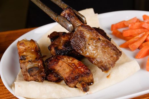 Sliced grilled pork ribs on pita bread with grilled carrots on a white plate on a wooden background.