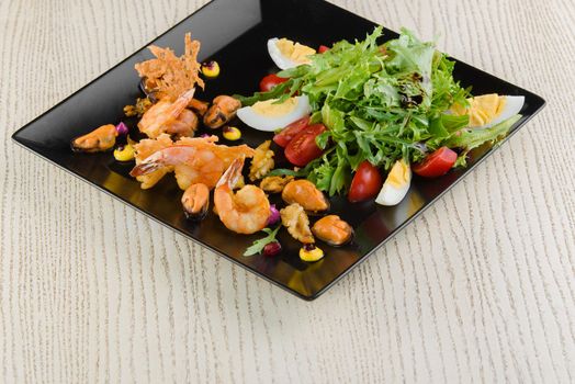Salad with shrimps, mussels, arugula, tomatoes, eggs, lettuce on a black square plate on a white wooden table.