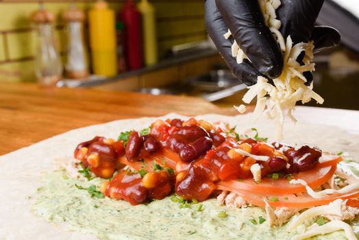 Burrito making process. To the meat, the chef adds cheese to beans and corn in tomato sauce, greens and tomatoes on pita with sauce. Mexican dish.