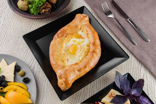 Khachapuri with cheese and tomatoes, egg, slices of vegetables, basil on a white wooden table. Serving. View from above