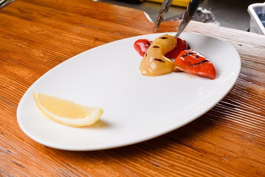 Grilled pepper and with lemon on a white plate on a wooden table. Serving the dish with metal tongs.