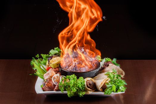 Flame meat in a clay bowl. Meat with fire on plate with grilled vegetables, kebab in pita bread decorated with herbs.