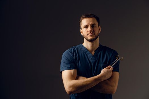 Handsome doctor with surgical scissors on dark background. Confident man holding medical equipment in hands and smiling. Happy male posing in studio