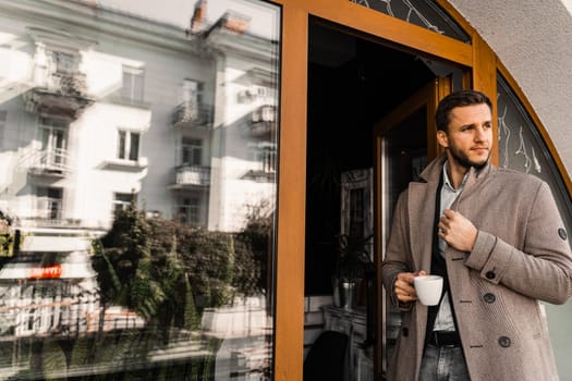 Handsome man with cup of coffee posing in coat in cafe. Male lifestyle