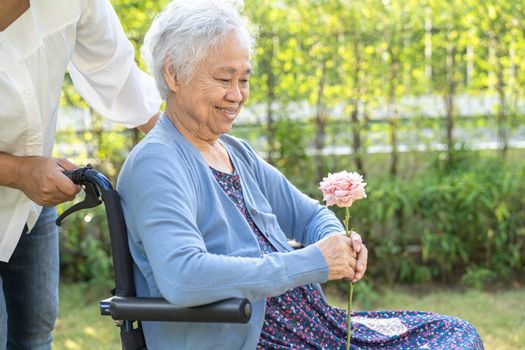 Caregiver daughter hug and help Asian senior or elderly old lady woman holding red rose on wheelchair in park.