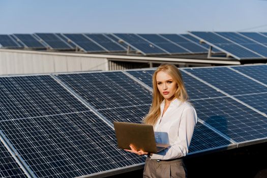 Investor woman stand with laptop near blue solar panels row on the ground. Free electricity for home. Sustainability of planet. Green energy.