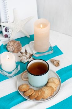 A study break: a hot mug of black tea with homemade cookies on a striped tablecloth; wax candles, decorative starfish, seashell, pile of notebooks on a white background.