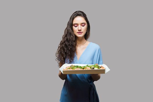 Girl courier with pinsa romana gourmet italian cuisine on grey background. Holding scrocchiarella traditional dish. Food delivery from pizzeria. Pinsa with meat, arugula, olives, cheese.