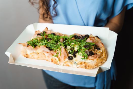 Pinsa romana in white delivery box. Courier holding crocchiarella gourmet italian cuisine on white background. Food delivery from pizzeria. Pinsa with meat, arugula, olives, cheese