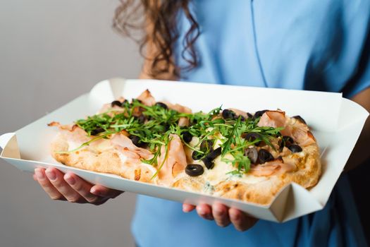 Pinsa romana in white delivery box. Courier holding crocchiarella gourmet italian cuisine on white background. Food delivery from pizzeria. Pinsa with meat, arugula, olives, cheese