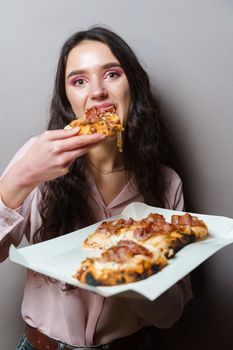 Girl courier eating pinsa pizza romana gourmet italian cuisine on grey background. Holding scrocchiarella traditional dish. Food delivery from pizzeria. Pinsa with meat, arugula, olives, cheese.
