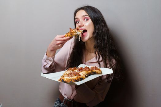 Girl courier eating pinsa pizza romana gourmet italian cuisine on grey background. Holding scrocchiarella traditional dish. Food delivery from pizzeria. Pinsa with meat, arugula, olives, cheese.