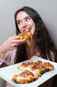 Girl courier eating pinsa pizza romana gourmet italian cuisine on grey background. Holding scrocchiarella traditional dish. Food delivery from pizzeria. Pinsa with meat, arugula, olives, cheese.