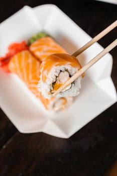 Holding philadelphia roll using chopstics for sushi. Roll with salmon, shrimp and mango. Japanese food delivery