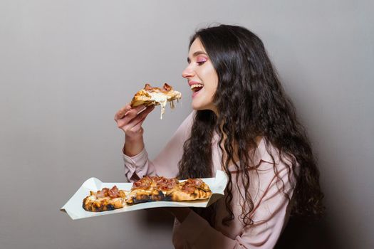 Girl courier eating pinsa pizza romana gourmet italian cuisine on grey background. Holding scrocchiarella traditional dish. Food delivery from pizzeria. Pinsa with meat, arugula, olives, cheese.