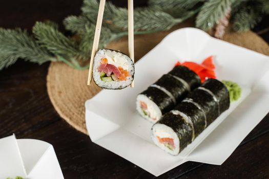Holding sushi roll using chopsticks on the food delivery container. Christmas decoration. Food delivery at new year eve. Roll with salmon, tuna, cucumber and soft cheese