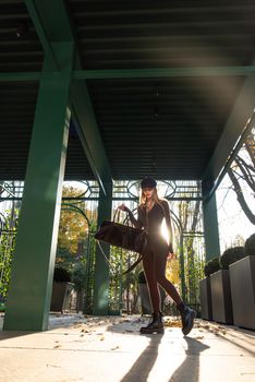 Fashionable young brunette woman with long legs. wearing solid long sleeve bodycon one piece jumpsuits posing with a leather brown travel bag on city street in old town