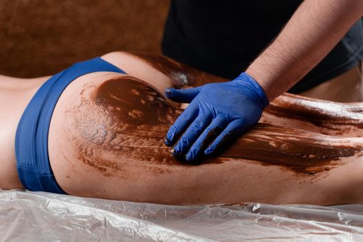 The masseur smears chocolate on the legs and thighs of the girl in the spa center. Beauty procedure chocolate wrap