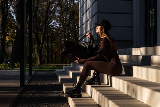 Fashionable young brunette woman with long legs. wearing solid long sleeve bodycon one piece jumpsuits posing with a leather brown travel bag on city street in old town