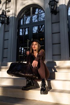 Fashionable young brunette woman with long legs. wearing solid long sleeve bodycon one piece jumpsuits posing with a leather brown travel bag on city street in old town