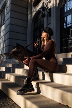 Fashionable young brunette woman with long legs. wearing solid long sleeve bodycon one piece jumpsuits posing with a leather brown travel bag on city street in old town